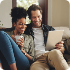 A young couple sit at home on the couch, reading from a tablet.  